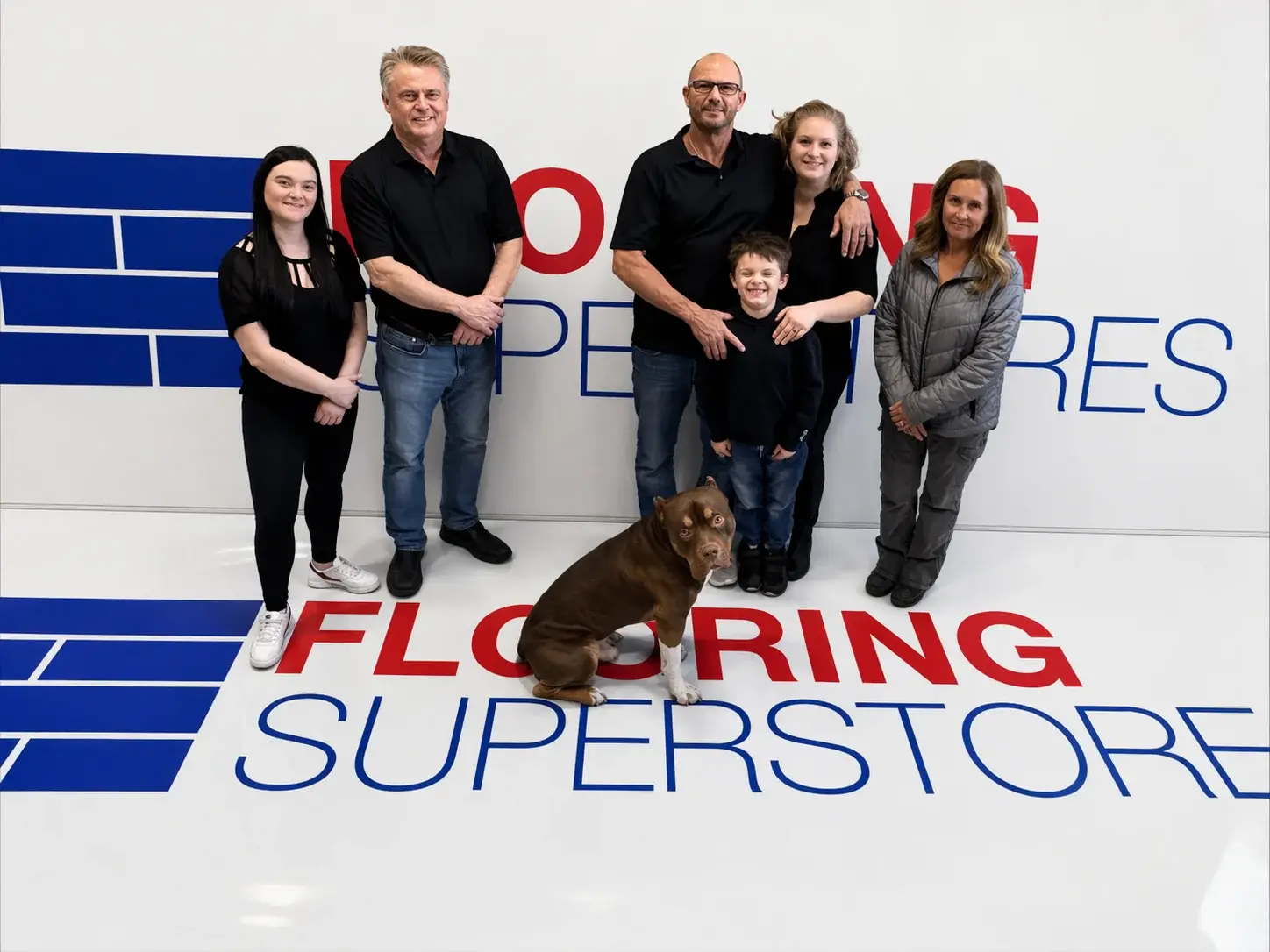 Kelowna Flooring Superstore team: showroom staff, installers, and shop dog in front of the Flooring Superstores mural
